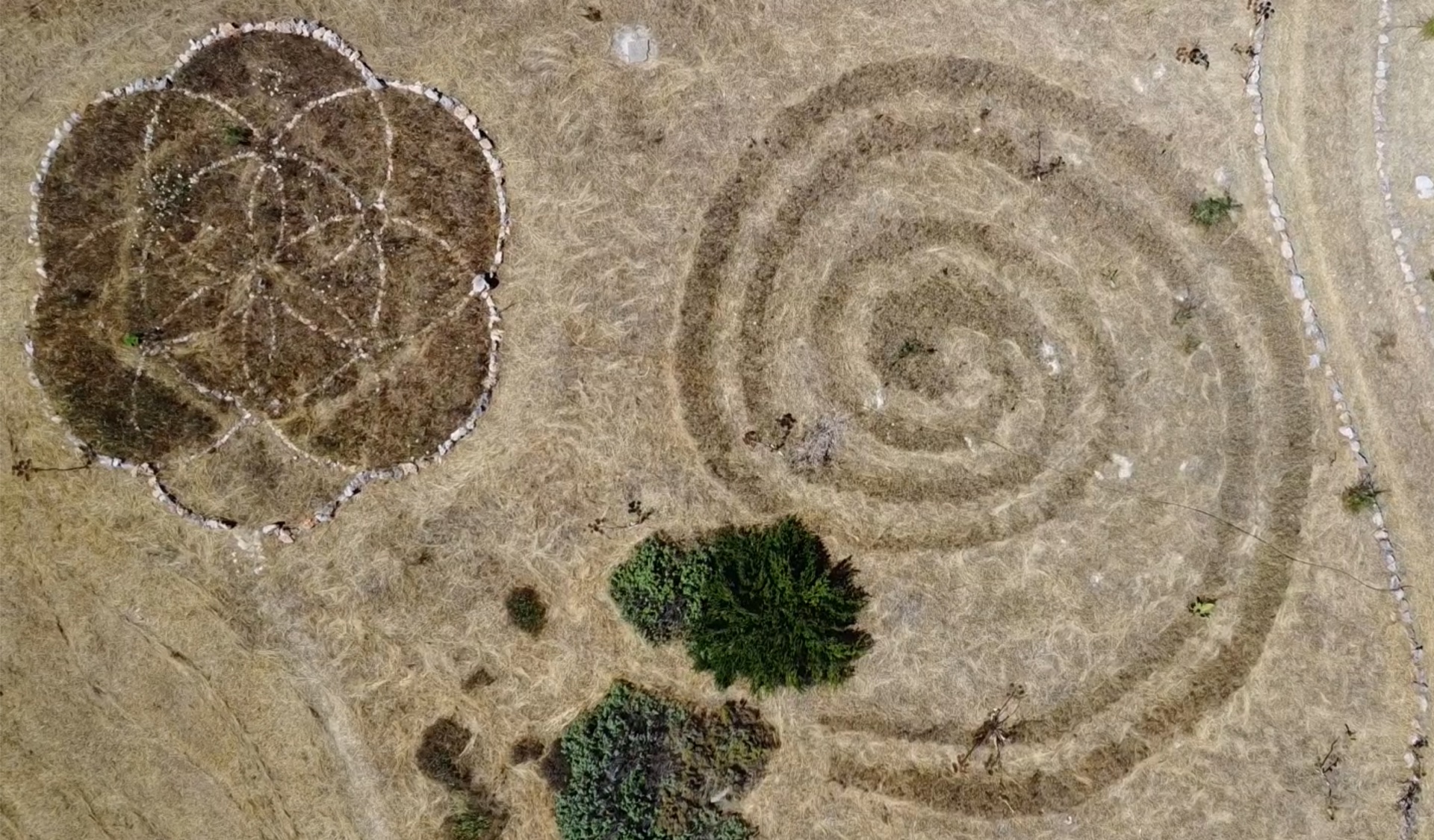 Una spirale tagliata nell’erba secca e un fiore della vita in pietre bianche, dall’alto